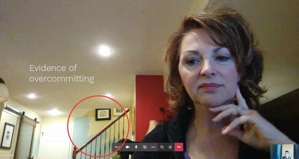 A woman sits in front of a computer during a video call. A red circle highlights a pile of items at the bottom of a staircase behind her, labeled Evidence of overcommitting.