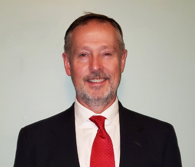 A middle-aged man with short hair and a beard is wearing a dark suit jacket, white dress shirt, and red tie. He is standing in front of a plain light-colored background and smiling.