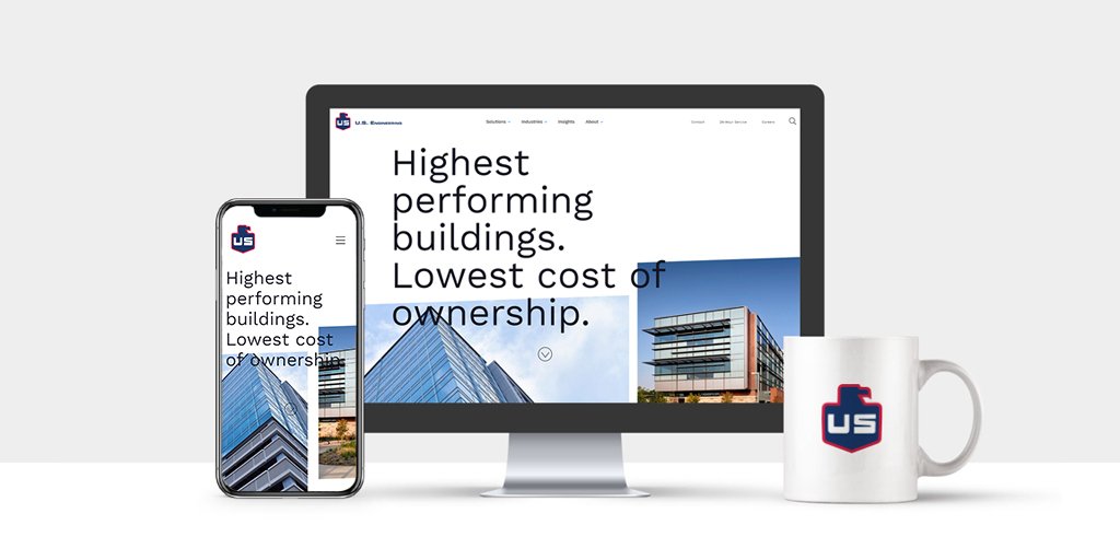 A smartphone, computer monitor, and white mug with a US logo are displayed side by side. The screens show a website with text: “Highest performing buildings. Lowest cost of ownership.” An office building is in the background.