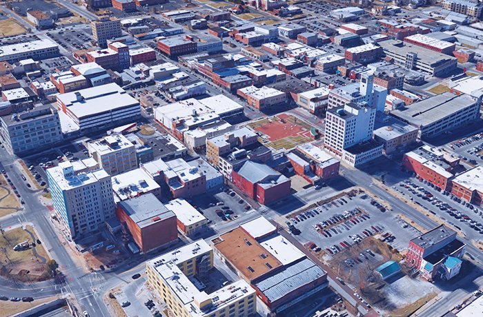 Aerial view of a city downtown area showing numerous buildings with flat rooftops, parking lots, streets laid out in a grid pattern, and parked cars. The surroundings include both commercial and residential structures.