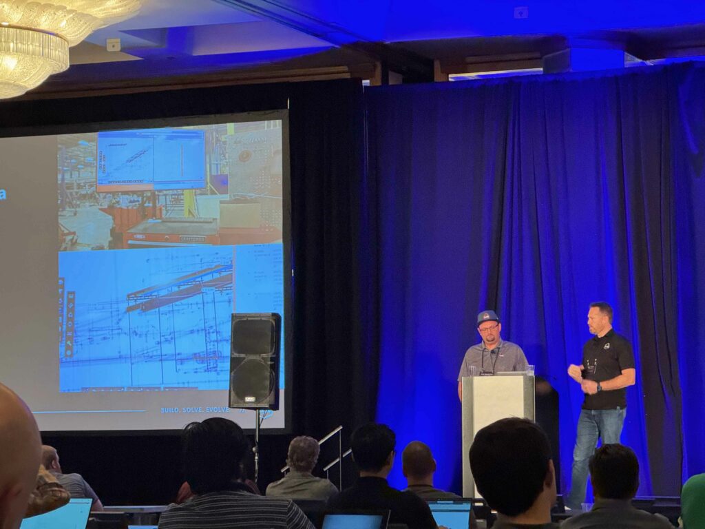 Two men stand on stage, one at a podium and one beside him with a microphone. A large screen displays diagrams and images behind them. Audience members sit facing the stage in a conference room.