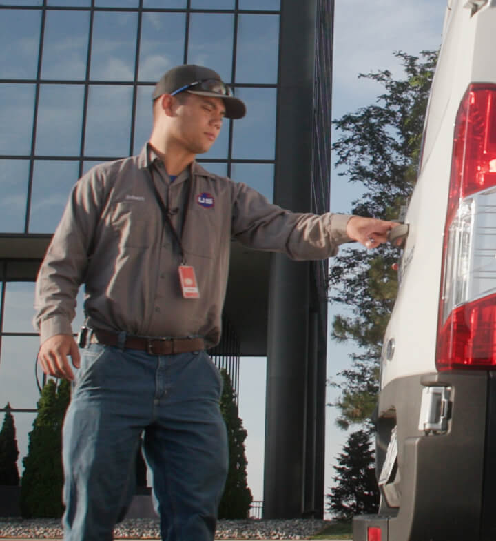 A man wearing a gray shirt, jeans, and a baseball cap stands outside near a white van, reaching toward the vehicle’s back door. A modern glass building and trees are visible in the background.
