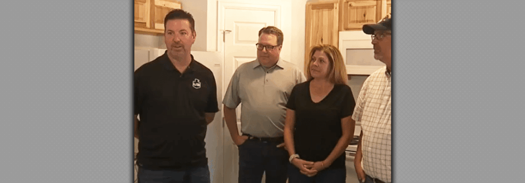 Four adults stand in a kitchen with light wooden cabinets; the man on the left is speaking while the other three people listen and look in his direction.