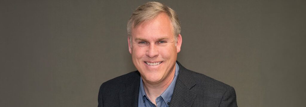 A middle-aged man with short, light hair is smiling, wearing a dark checked blazer over a blue shirt, against a plain grey background.