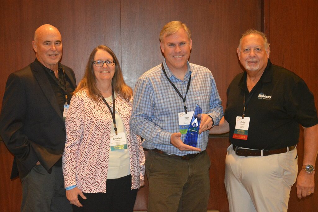 Four adults stand indoors in front of a wooden wall. One person in the center holds a blue award. All are wearing conference name badges and are smiling at the camera.