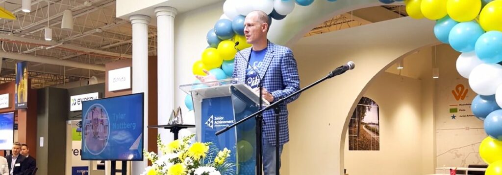 A man in a blue checked jacket speaks at a podium decorated with flowers, under an arch of yellow, blue, and white balloons. A screen and microphone stand are nearby in a modern indoor event setting.