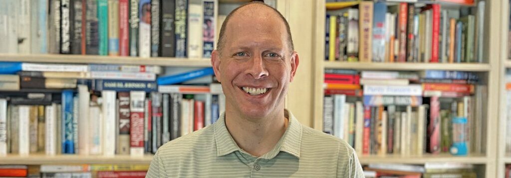 A man wearing a light green striped polo shirt smiles at the camera while standing in front of a bookshelf filled with various books.