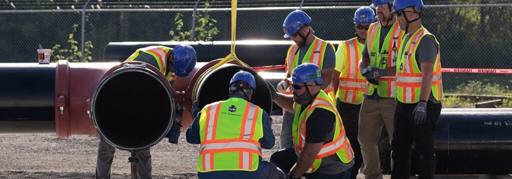Six construction workers wearing safety vests and blue helmets work together by large black pipes at an outdoor site. One worker holds a yellow lifting strap attached to a pipe section. A chain-link fence is in the background.
