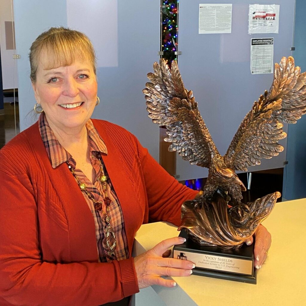 A woman with light brown hair in a ponytail, wearing a red cardigan and patterned blouse, stands indoors smiling and holding a large bronze eagle statue on a base with an engraved plaque.