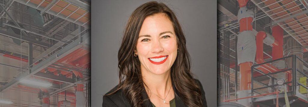 Woman with long dark hair and red lipstick, wearing a black blazer, smiles at the camera. The background features industrial equipment and pipes in a factory setting, partially blurred.