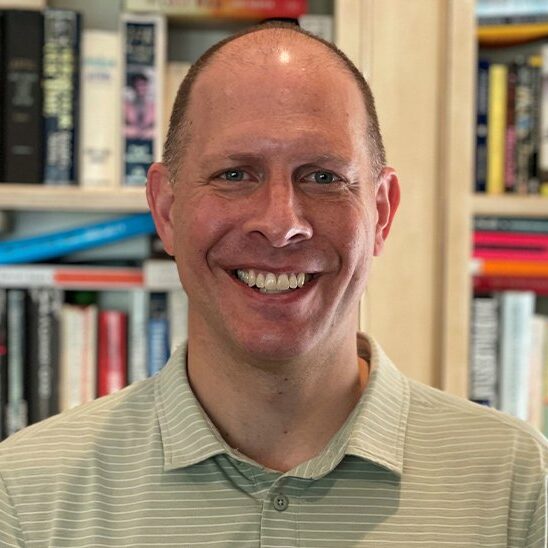 A man with a shaved head and a light green striped polo shirt smiles in front of a bookshelf filled with various books.