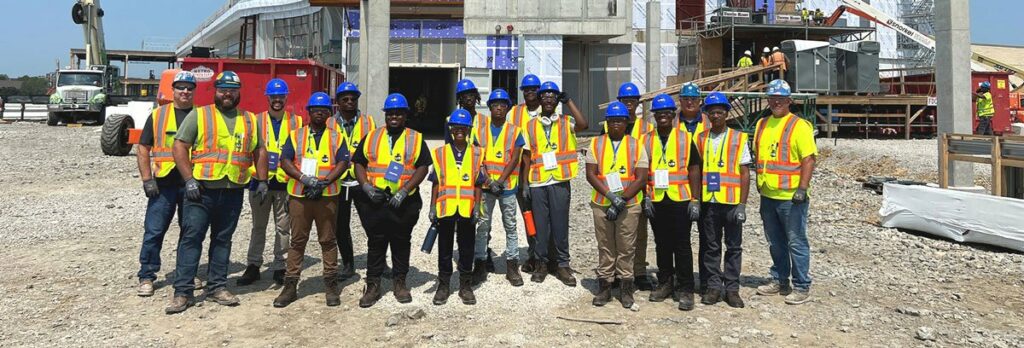 A group of construction workers and staff wearing safety vests and helmets stand in a line at a construction site with unfinished buildings and equipment in the background.