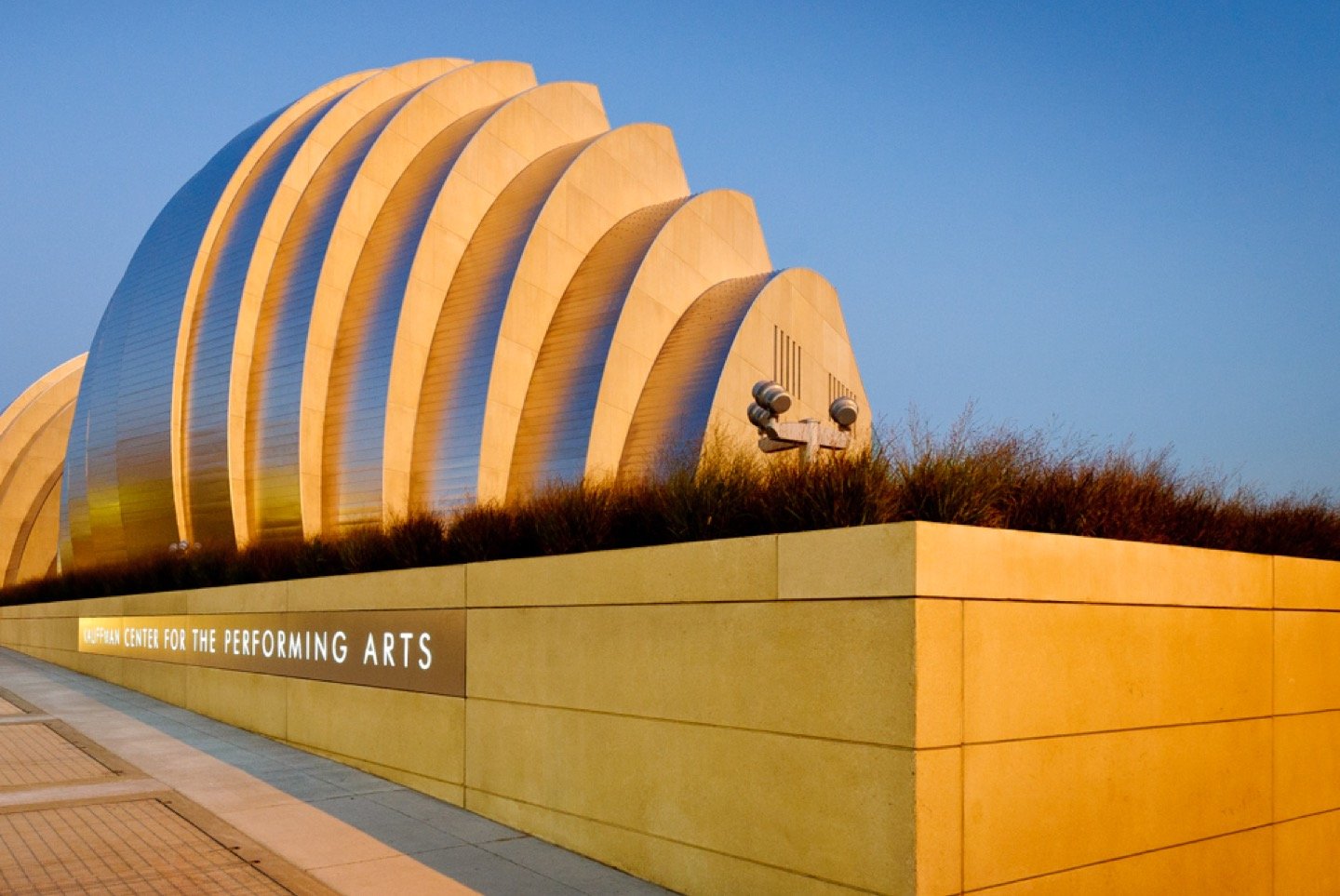 A modern building with curved, overlapping metallic segments; the sign on a stone wall in front reads CENTER FOR THE PERFORMING ARTS. The structure is lit by sunlight under a clear blue sky.