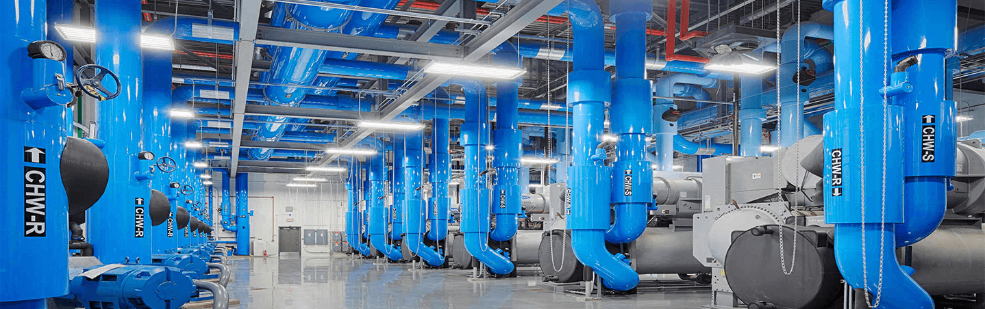 A large industrial facility with many bright blue pipes and valves labeled “CHWR” and “CHWS,” overhead lighting, and metal machinery lined up throughout the clean, spacious room.