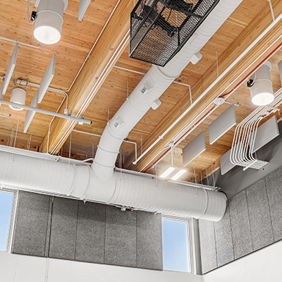 Exposed ceiling with wooden panels, white ventilation ducts, hanging light fixtures, acoustic panels, and electrical conduits in a modern interior space.