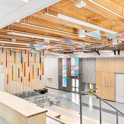 A modern, open interior space with light wood finishes, exposed beams, colorful wall art, geometric decorations hanging from the ceiling, large windows, and a staircase leading to a seating area.