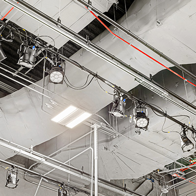 Ceiling view of a studio or stage showing multiple mounted lights, cables, pipes, and ducts, with some fluorescent panels and metal beams visible.