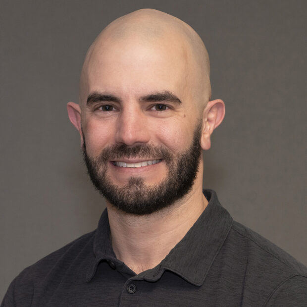 A man with a bald head and a dark beard smiles at the camera. He is wearing a dark collared shirt and is posed against a plain, neutral background.