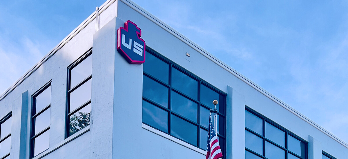 A white, two-story building with large glass windows is shown. The red and white US logo is mounted on the corner, and an American flag is visible in front of the building against a blue sky.