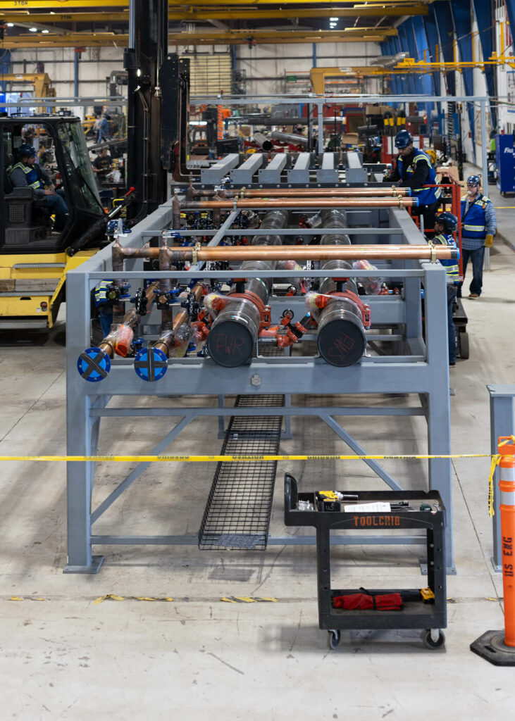 Industrial warehouse with large metal framework and pipes in the center, workers in safety gear nearby, a yellow forklift on the left, and a rolling tool cart in the foreground. The area is sectioned off with caution tape.