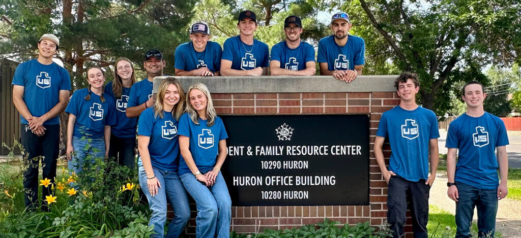 A group of twelve people wearing matching blue shirts pose together outside in front of a sign for the Parent & Family Resource Center and Huron Office Building at 10290 Huron. Some are sitting, others are standing.