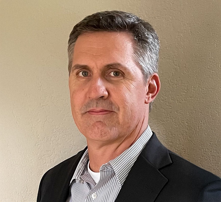 A man wearing a dark blazer over a light striped shirt stands in front of a plain, textured wall, looking directly at the camera with a neutral expression.