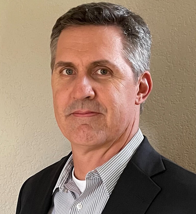 A middle-aged man with short gray hair wearing a dark blazer over a light striped shirt stands in front of a beige textured wall, looking slightly to the left of the camera.