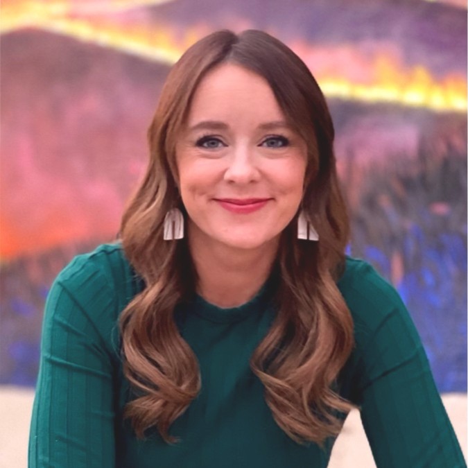 A woman with long, wavy brown hair and light skin is smiling at the camera. She is wearing a green top and white earrings. The background is a blurred, colorful mural with shades of purple, pink, and yellow.