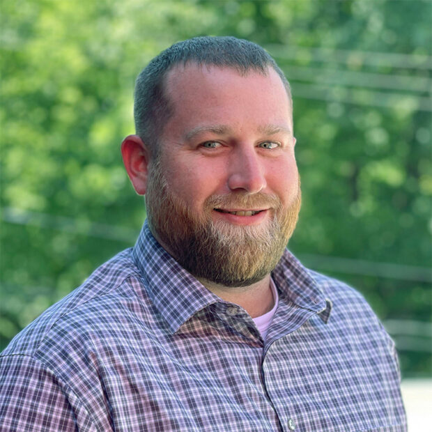 A man with a short beard and short hair, wearing a plaid button-up shirt, stands outdoors in front of a blurred background of green trees. He is smiling and facing the camera.
