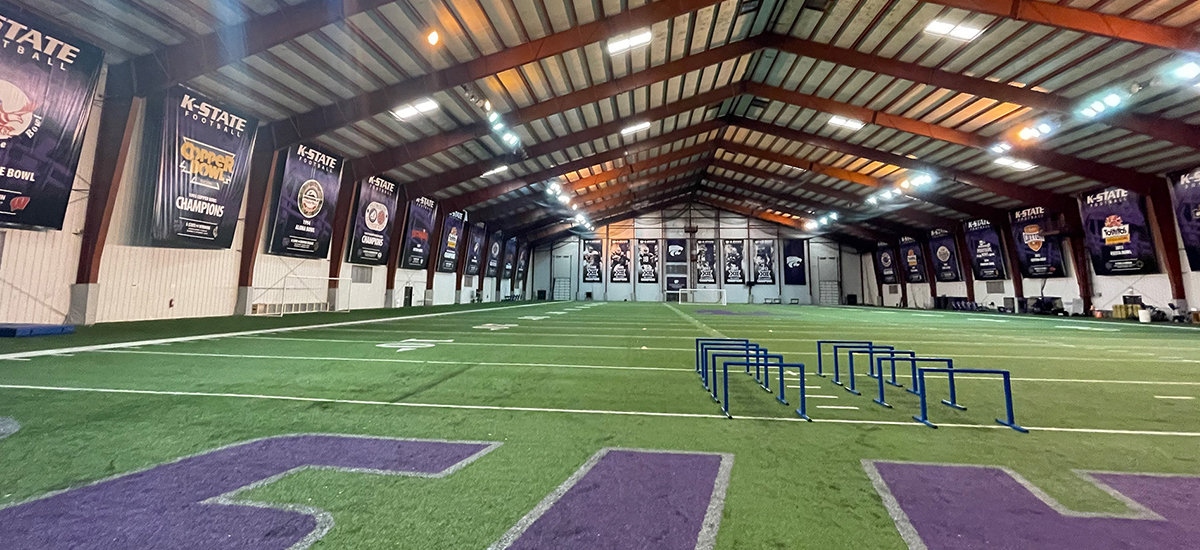 An indoor football practice facility with artificial turf, blue training hurdles, and large championship banners hanging on the walls under a metal roof. The space is well-lit and spacious.