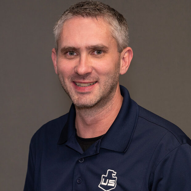 A man with short gray hair and light skin is smiling at the camera. He is wearing a navy blue collared shirt with a white logo on the left side, ideal for construction or offsite fabrication work. The background is plain and gray.