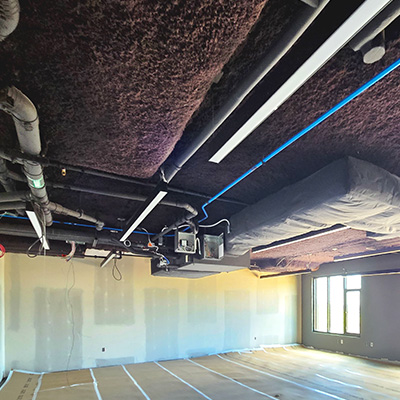 A room under construction with exposed pipes, ducts, and wiring on the ceiling, unfinished walls, and protective covering on the floor. Sunlight enters through a window on the right side.