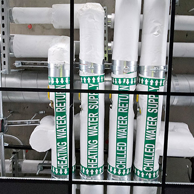 Several insulated pipes are labeled HEATING WATER RETURN, HEATING WATER SUPPLY, and CHILLED WATER RETURN with green and white text and arrows, running vertically along an industrial wall.