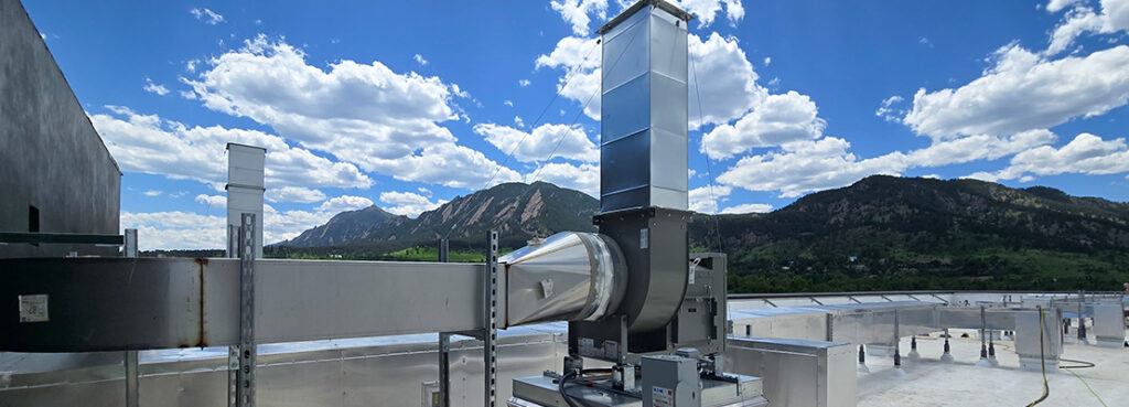 A large industrial ventilation system is installed on a rooftop with mountains and a blue sky with scattered clouds in the background. Various metal ducts and units are visible around the main equipment.