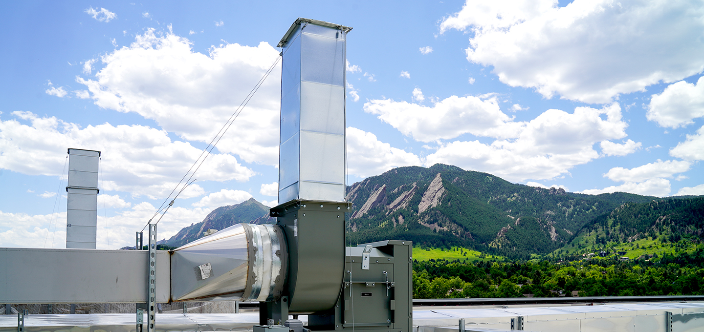 A large industrial ventilation system is mounted on a rooftop with green mountains and a partly cloudy sky in the background.