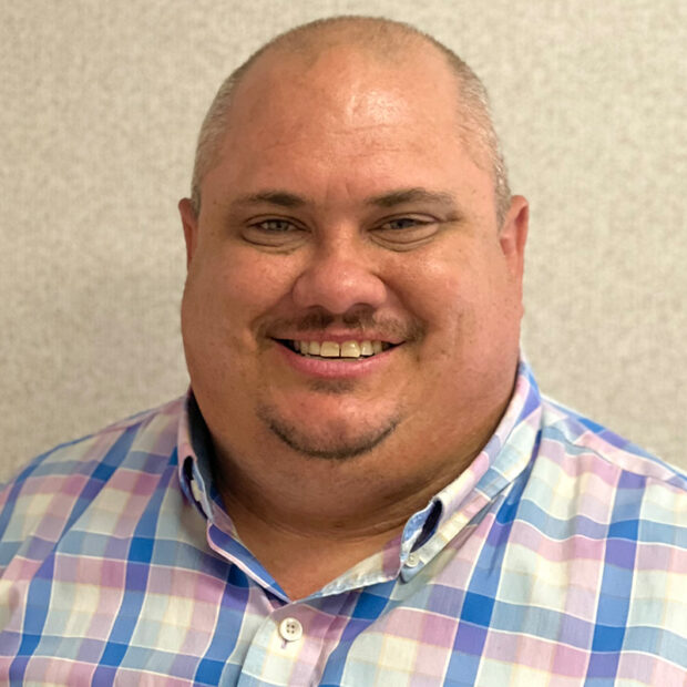 A man with short-cropped hair and a goatee is smiling at the camera. He is wearing a pastel plaid button-up shirt and is posed in front of a plain, light-colored background.
