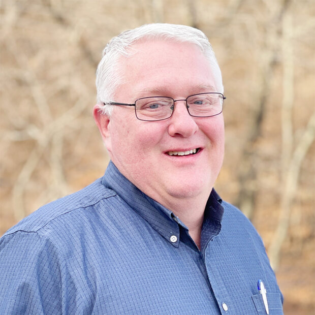 A middle-aged man with short gray hair and glasses is smiling. He is wearing a blue button-up shirt with pens in the pocket. The background is outdoor with trees and soft natural light.