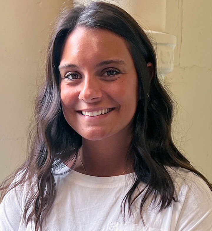 A woman with long, wavy brown hair and medium skin tone is smiling at the camera. She is wearing a white top and is positioned in front of a light-colored wall.