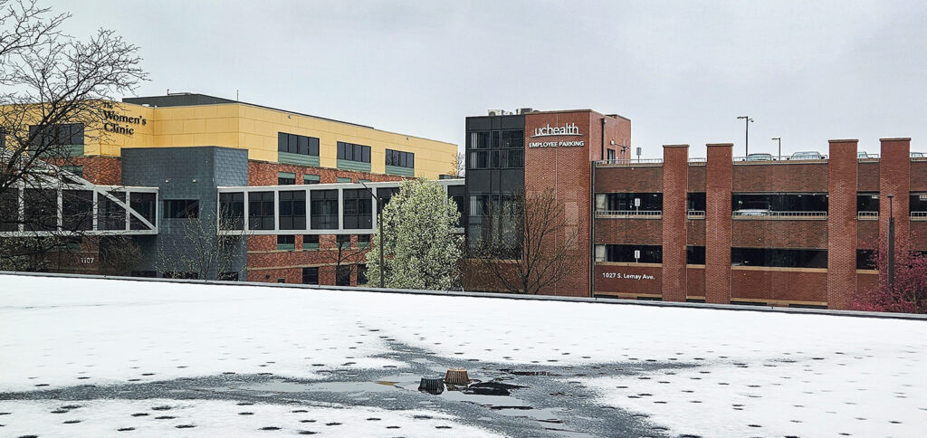 A hospital complex with brick and yellow buildings, a skywalk connecting structures, and a parking garage. The foreground shows a flat roof partially covered in melting snow under an overcast sky.