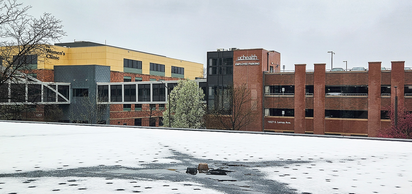 A hospital complex with brick and yellow buildings, a skywalk connecting structures, and a parking garage. The foreground shows a flat roof partially covered in melting snow under an overcast sky.
