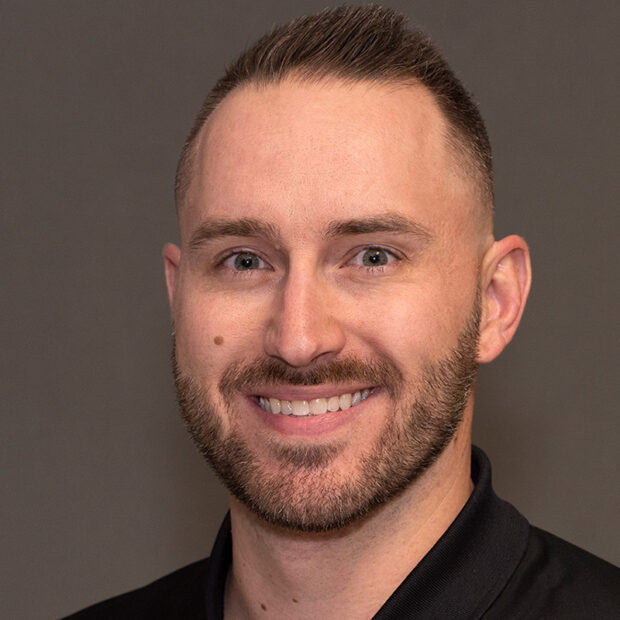 A man with short brown hair and a trimmed beard is smiling at the camera. He is wearing a black collared shirt and is posed in front of a plain, neutral background.
