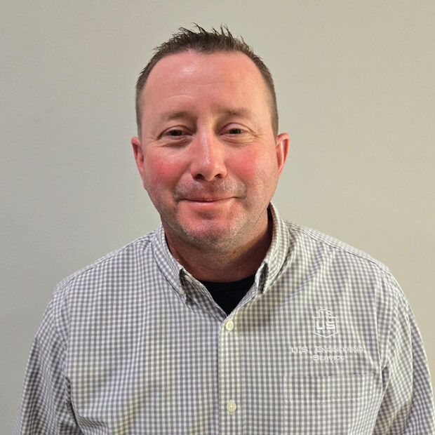 A man with short brown hair wearing a gray and white checkered button-up shirt stands in front of a plain light-colored wall. The shirt has a logo and text that reads “USI Insurance Services.”.