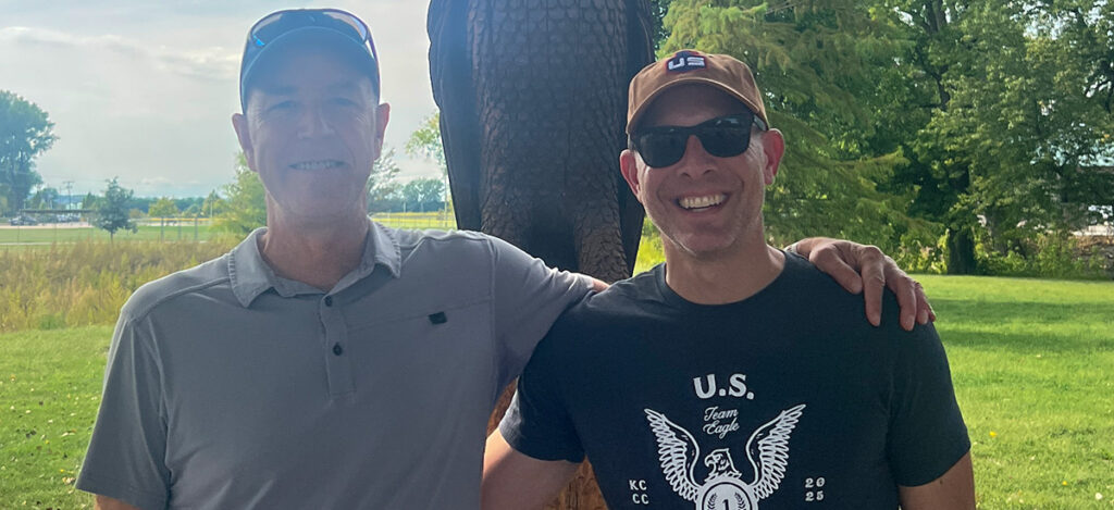 Two men standing outdoors on a sunny day, smiling at the camera. One is wearing a light gray shirt and baseball cap; the other wears sunglasses, a brown cap, and a black “U.S. Team Eagle” shirt. Green trees and grass are in the background.