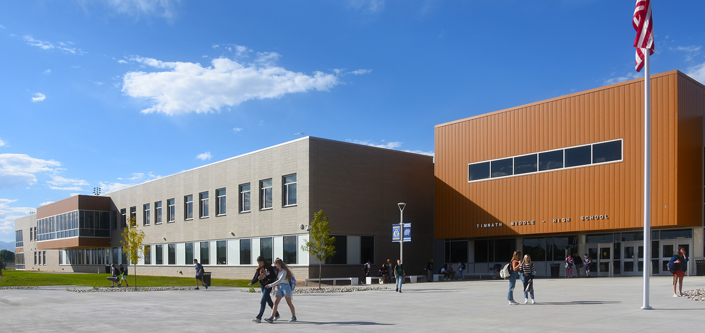 A modern two-story school building with large windows and an orange accent section; students walk and gather outside on a sunny day, and an American flag stands on a pole in the foreground.