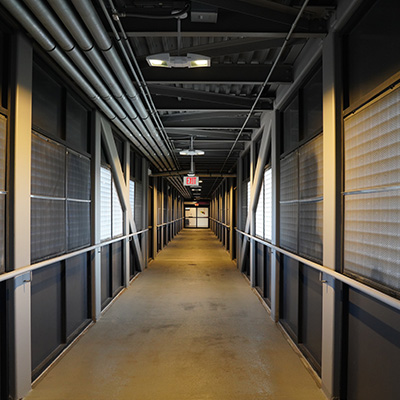 A long, industrial-style hallway with metal beams, overhead pipes, grated windows on both sides, and a concrete floor, illuminated by ceiling lights. An exit sign is visible at the far end.