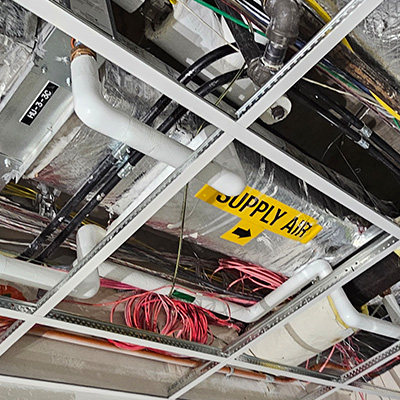 Exposed ceiling with white pipes, pink electrical cables, and insulation. A yellow “SUPPLY AIR” sign with a black arrow is visible among the various mechanical and electrical components. Ceiling tiles are partially removed.