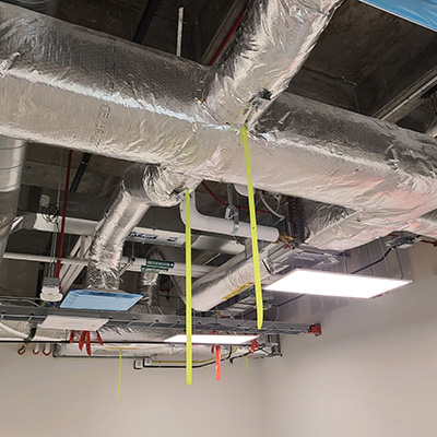 Exposed HVAC ductwork wrapped in insulation, with yellow and red straps hanging down from the pipes. The ceiling is unfinished and there are fluorescent lights and electrical conduits visible.