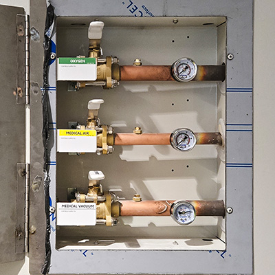 A wall-mounted medical gas panel with three labeled valves: oxygen, medical air, and medical vacuum. Each valve has a copper pipe and a pressure gauge. The panel is open, exposing the components.