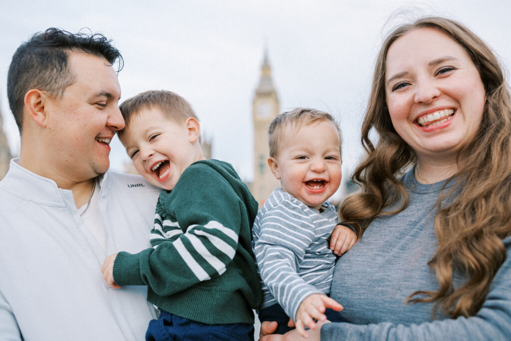 A man and woman smile while holding two young children who are also smiling. Big Ben is visible in the background, indicating they are in London. The group appears to be enjoying a day out together.