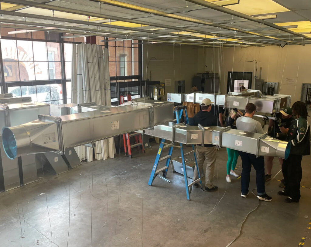 A group of people stand around a large metal air duct system in an industrial workshop. The room has concrete floors, fluorescent lights, and various construction materials along the walls.